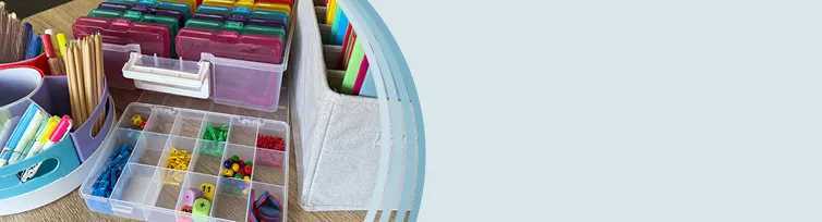 Auf einen Tisch steht eine transparente Sortierbox mit Würfeln, Klammern und Perlen darin, daneben steht ein Tisch-Organizer mit Stiften, Kleber und Scheren