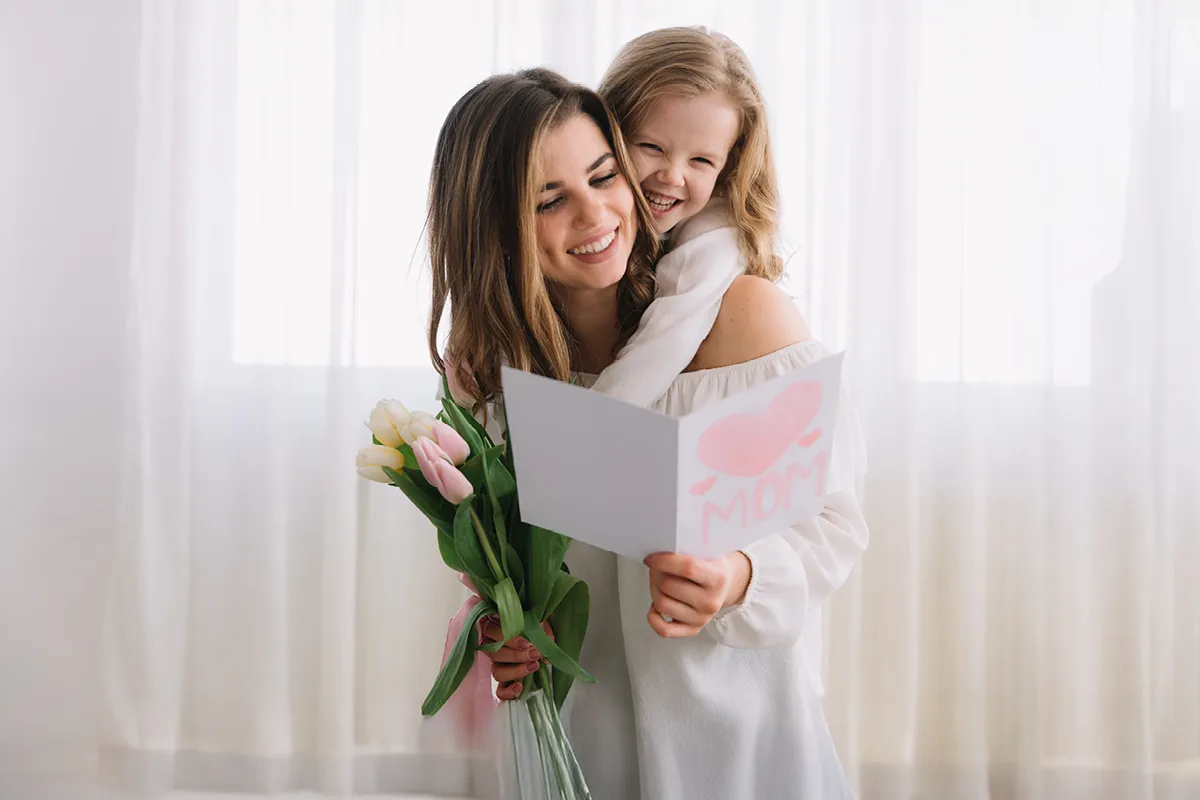 Glückliche Mutter mit Kind auf dem Arm und hat selbstgebastelte Karte zum Muttertag in der Hand sowie ein Strauß Tulpen