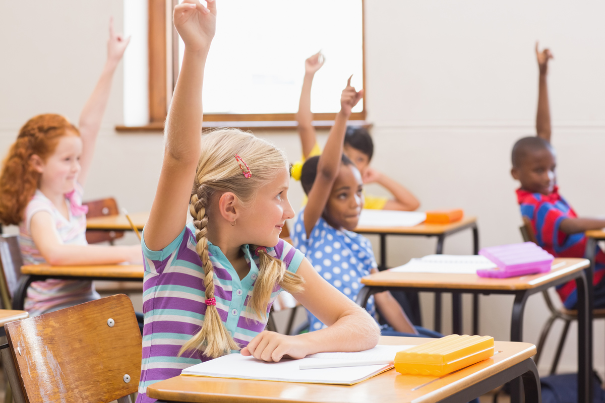Grundschulkinder sitzen im Klassenzimmer an Tischen und melden sich