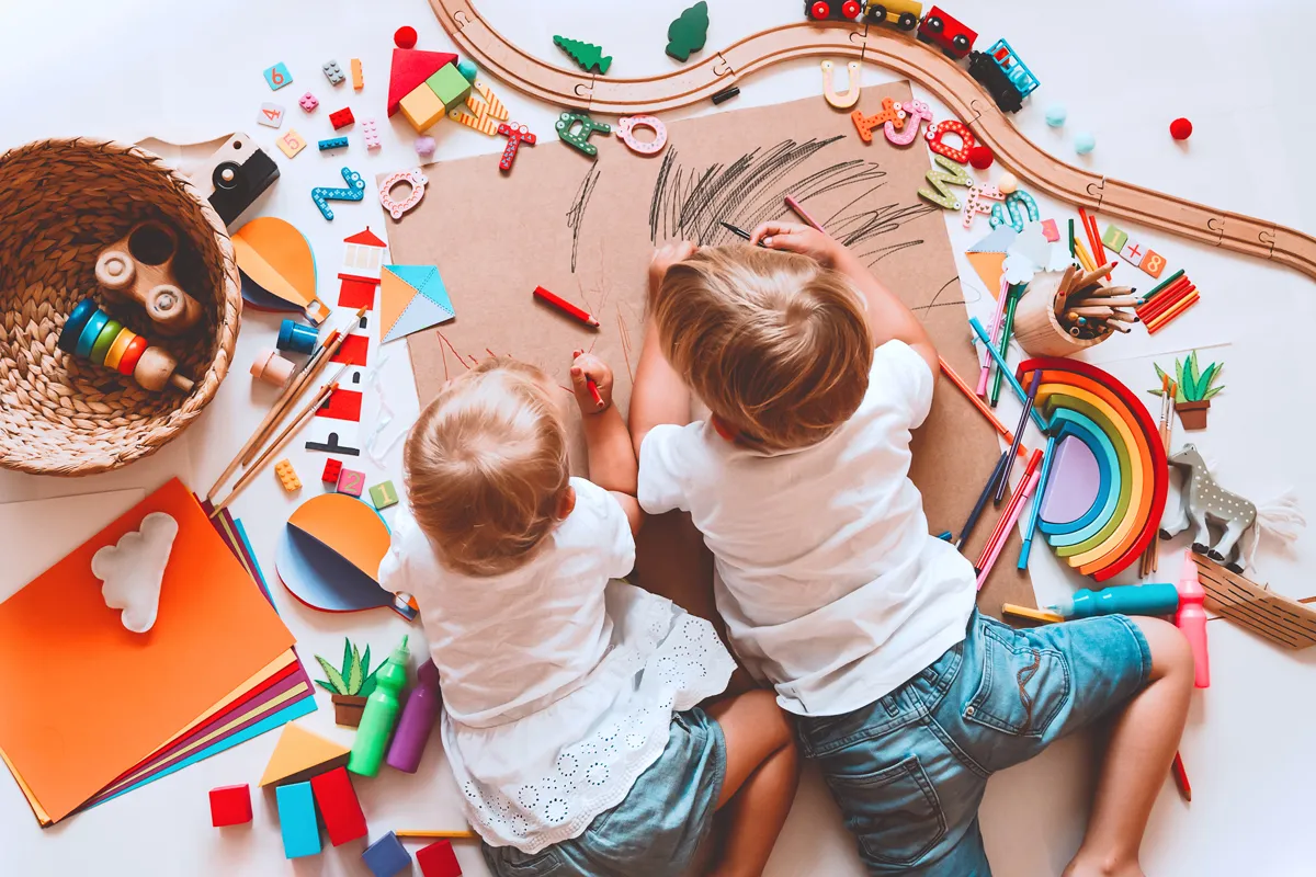 Von oben fotografierte zwei Kleinkinder beim gemeinsamen Malen auf großem Papier, umringt von Holzspielzeug, Bausteinen, Buchstaben und Farben