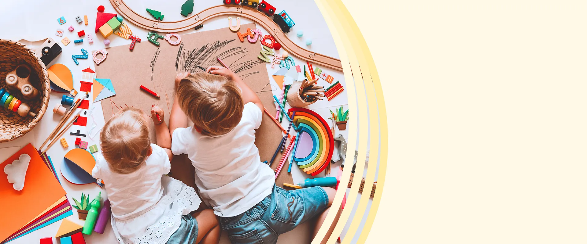 Von oben fotografierte zwei Kleinkinder beim gemeinsamen Malen auf großem Papier, umringt von Holzspielzeug, Bausteinen, Buchstaben und Farben