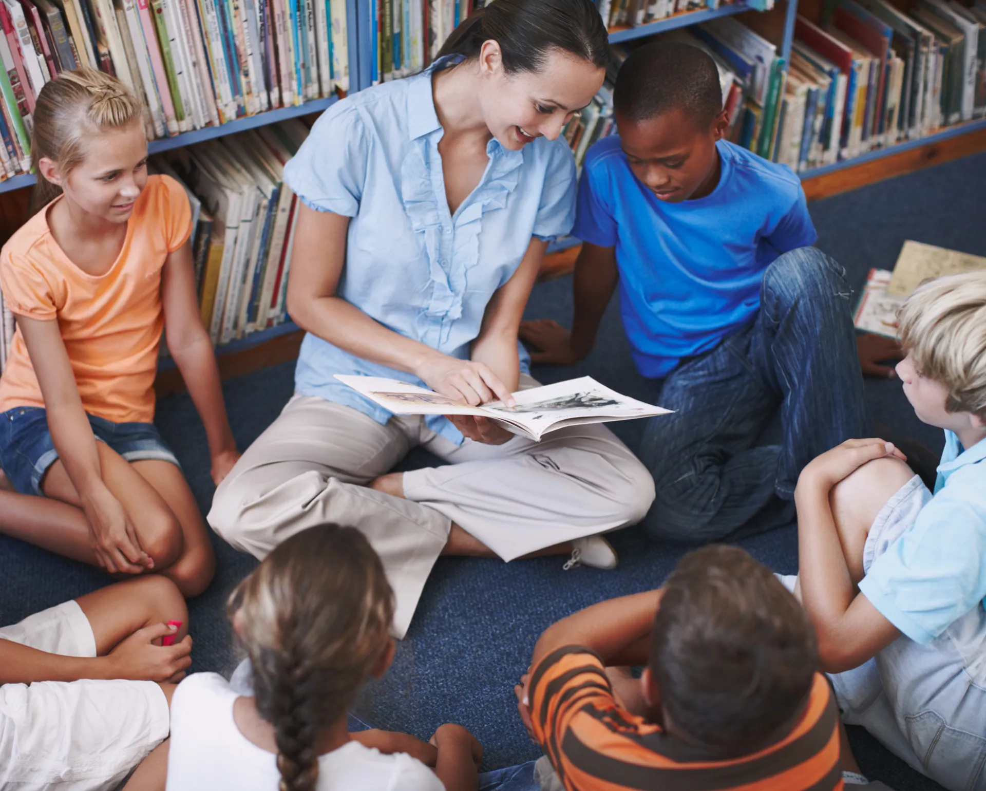 Lehrerin sitzt im Kreis mit Schülern in der Bibliothek und hält ein Buch in der Hand