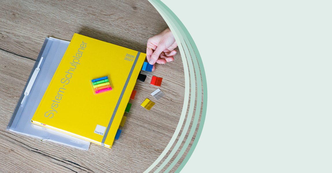 Auf einem Holztisch liegen ein gelber System-Schulplaner mit farbigen Register-Taben und ein Markierungsstreifen Set