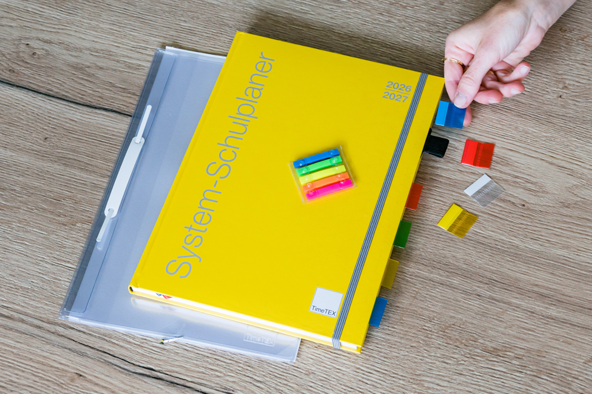 Auf einem Holztisch liegen ein gelber System-Schulplaner mit farbigen Register-Taben und ein Markierungsstreifen Set