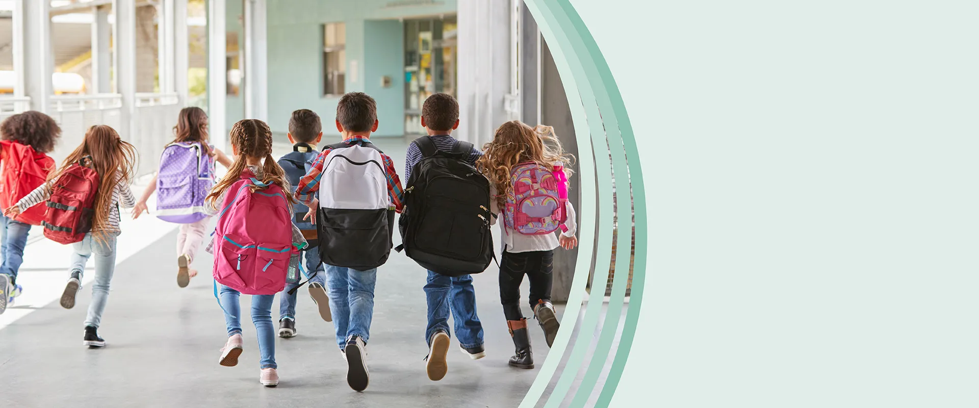 Gruppe von Schulkindern mit bunten Rucksäcken wird von hinten fotografiert, wie sie gemeinsam durch einen sonnendurchfluteten Schulflur rennen
