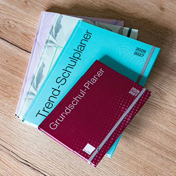 Stapel mit unterschiedlichen TimeTEX Planer wie den Grundschul-Planer und den Trend-Schulplaner liegen auf einem Holztisch leicht gefächert
