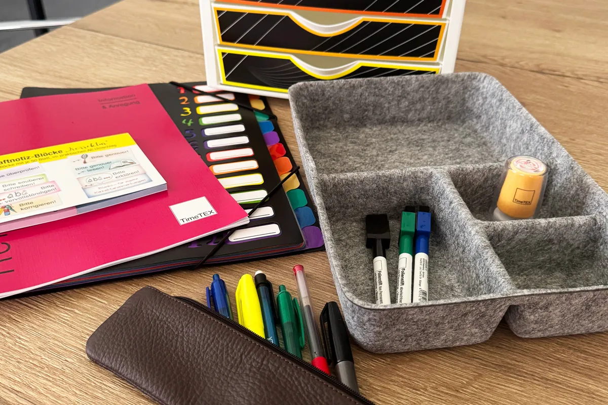 Auf einem Tisch liegen Planer, eine Fächermappe, Haftnotizen und eine graue Organisator-Box aus Filz mit Stiften und Stempeln